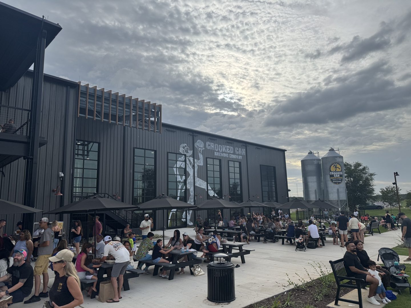 The Crooked Can Brewing Company building in Minneola, Florida, at dusk on its first Saturday open. A two-story dark metal warehouse carries a painted figure of a man tipping a barrel beside the words CROOKED CAN BREWING COMPANY. Black patio umbrellas and picnic tables fill the concrete apron in front, occupied by roughly forty people. Two galvanized grain silos stand at the right of the frame under a broken cloud deck.