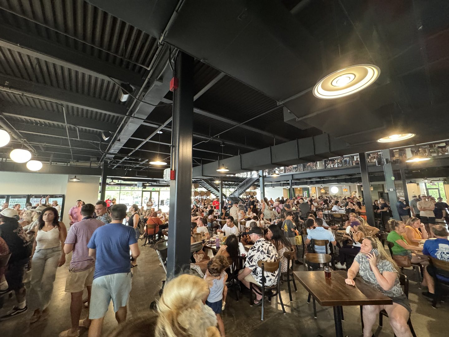 The interior of the City Center Market food hall at Crooked Can Brewing in Minneola, Florida, on its first Saturday open. Several hundred people fill communal tables across a two-level industrial room with exposed black ductwork, steel columns, and disc pendant lights. A mezzanine walkway carrying more diners runs along the far wall.