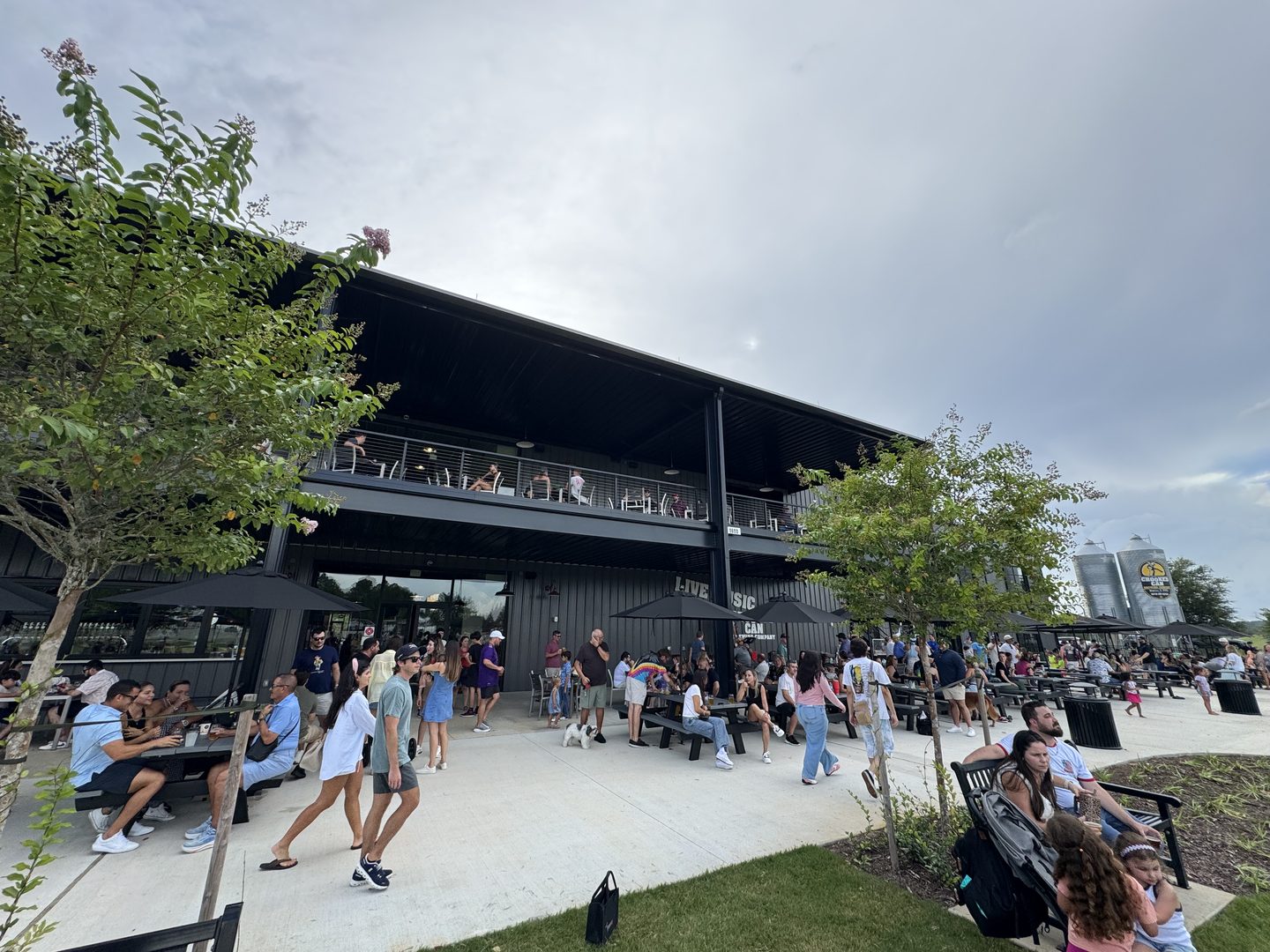 The patio and second-floor deck at Crooked Can Brewing in Minneola, Florida, on its first Saturday open. People fill the covered upper balcony and the picnic tables below; a queue runs along the building's open frontage beneath a LIVE MUSIC sign. Two branded grain silos stand at the far right beyond a row of black umbrellas.