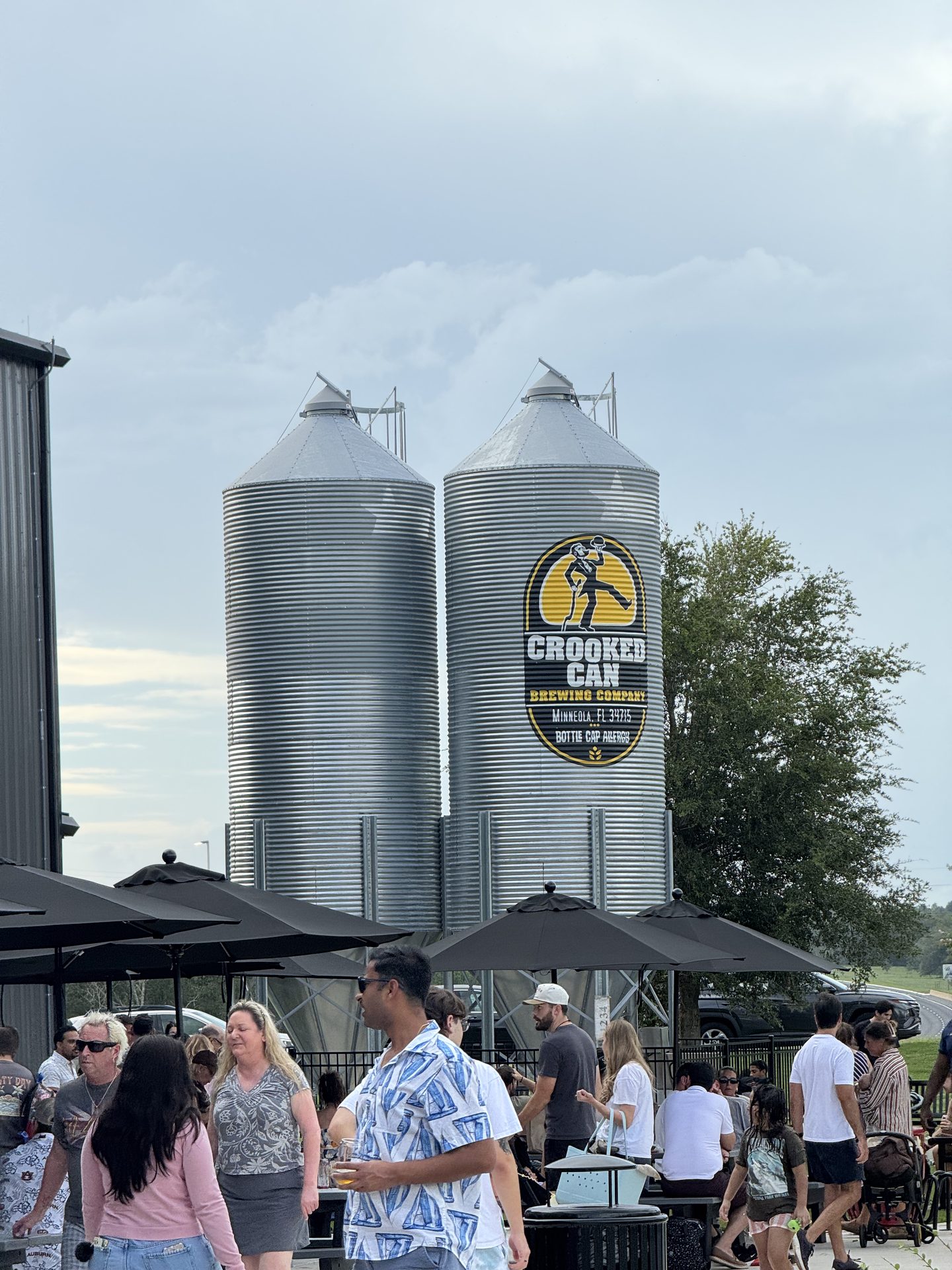 Two galvanized grain silos at Crooked Can Brewing in Minneola, Florida. The right-hand tank carries the company's oval logo, a figure tipping a barrel, above the words CROOKED CAN BREWING COMPANY, MINNEOLA, FL 34715, BOTTLE CAP RANCH. Black patio umbrellas and a crowd of visitors occupy the foreground.